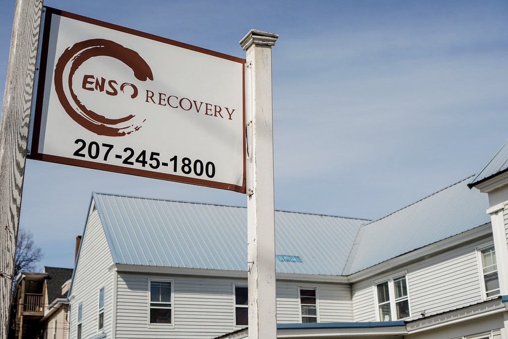 Enso Recovery sign with the persimmon ensō circle and the 207-245-1800 admissions phone number outside the Augusta clinic, white clapboard building under a soft Maine sky
